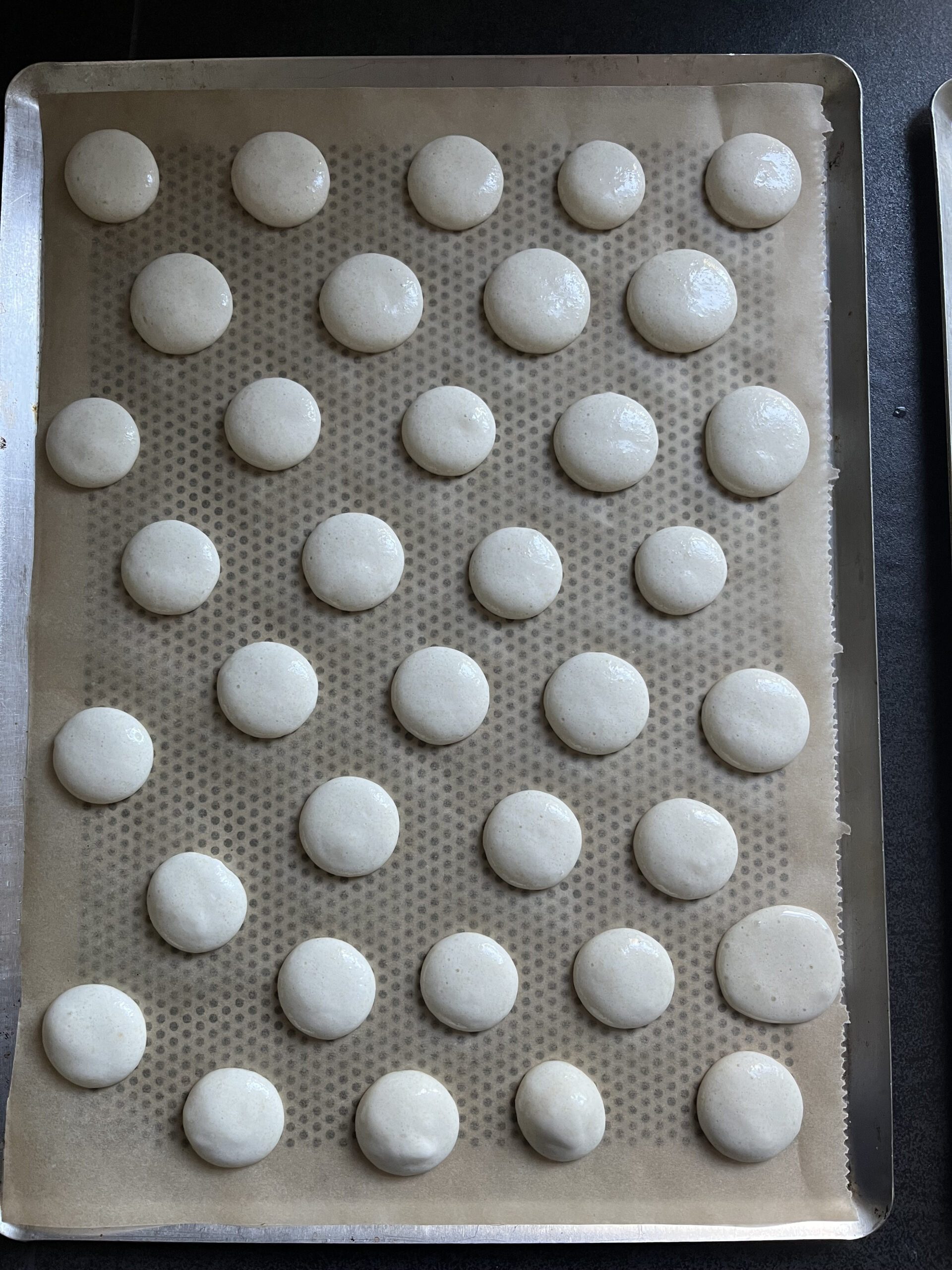 Kleine runde Macarons auf ein mit Backpapier belegtes Lochblech spritzen, sie dann sofort mit Wasser besprühen